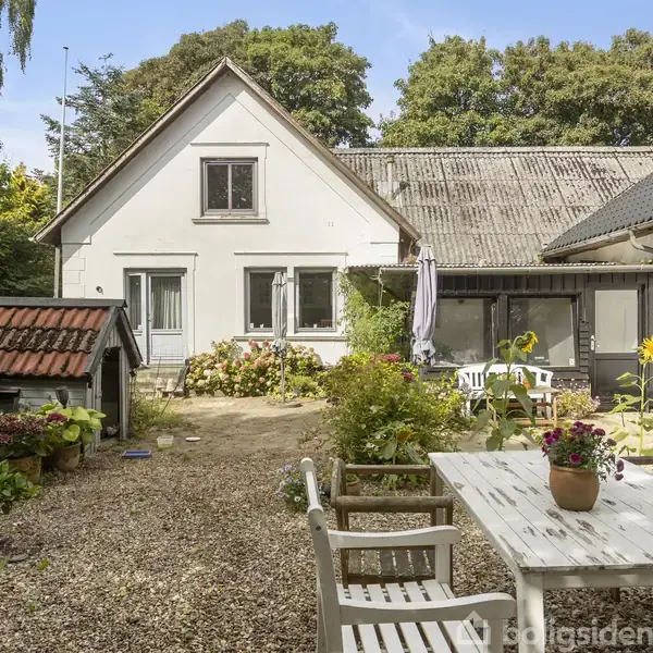 Hus med hvid facade og sadeltag. Foran huset er der en gårdsplads med et hvidt havebord, stole og blomsterkrukker. Omgivelserne har træer og en lille bygning med rødt tag.