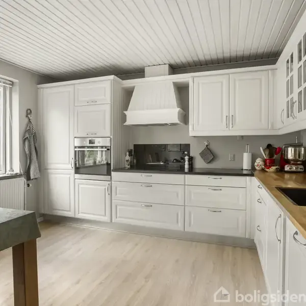 Lyse køkkenskabe med integrerede hvidevarer og emhætte; køkkenbord i træ til højre med vask; stort vindue mod haven; spisebord til venstre; lyst trægulv.