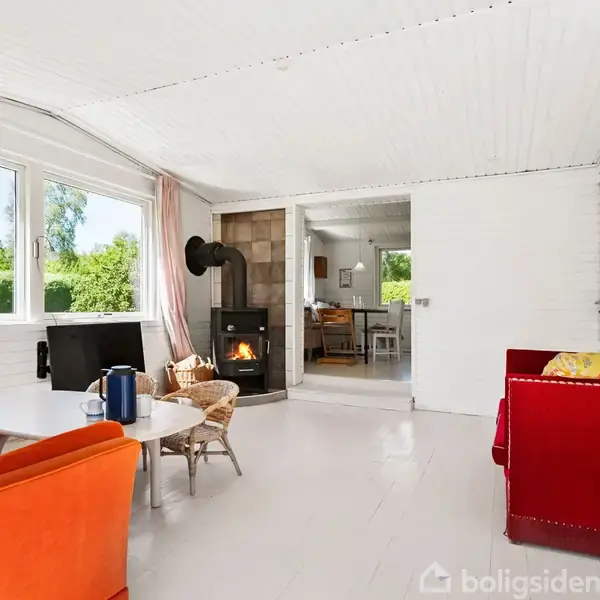 En hyggelig stue med en orange sofa, rød lænestol og et rundt bord. Brændeovn i hjørnet ved siden af en åbning, der fører til et køkken. Store vinduer giver udsigt til grønne planter udenfor.