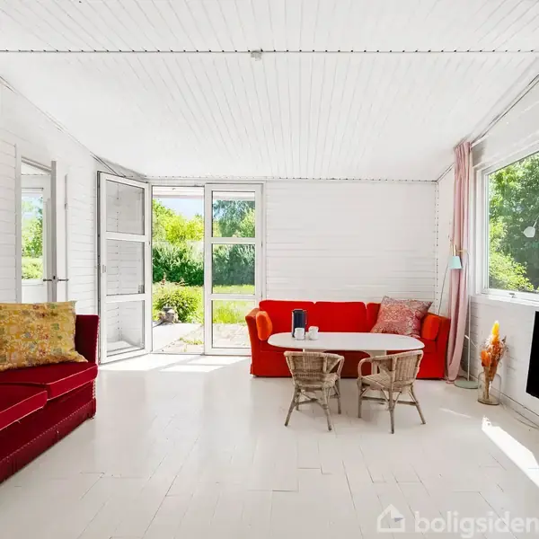 En lys stue indeholder to sofaer, hvoraf den ene er rød, placeret ved et rundt bord med stole. Stuen har store vinduer og en dør, der åbner ud til en have.