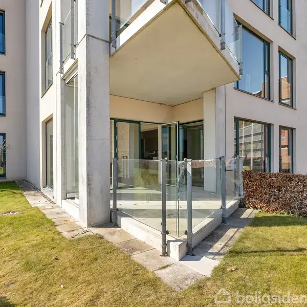 En balkon med glasrækværk er åben til en velplejet have. Bygningen har store vinduer og ligger i et moderne boligområde med grå pavilloner og brune buske.
