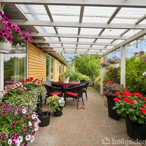En overdækket terrasse fyldt med blomsterkrukker i forskellige farver. Et bord med stole står midt i området, omgivet af tæt grøn beplantning og hængende blomsterkurve.