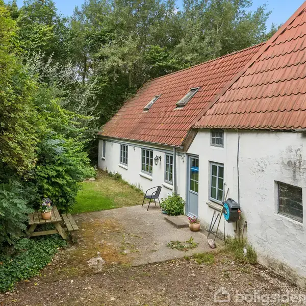 Et hvidt, langstrakt hus med rødt tegltag står stille omgivet af tæt, grøn bevoksning. En lille terrasse med møbler er placeret ved siden af husets hoveddør.