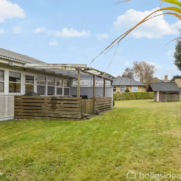 Hus med overdækket terrasse, omgivet af velplejet græsplæne i boligområde. Stenmur og andre huse i baggrunden kan ses, himlen er let overskyet.