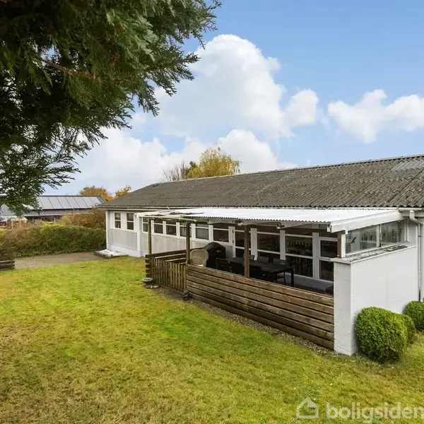En lang, hvid bygning med store vinduer har en lukket terrasse. Bygningen står på en velplejet græsplæne med buske og træer i baggrunden under en blå himmel.