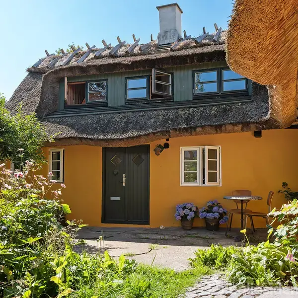 Stråtækt hus med åbne vinduer, omgivet af grønne planter og blomster. En lille terrasse med bord og stole ses, alt under en klar blå himmel.