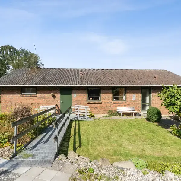Et rødt murstenshus med fladt tag står i en have med græsplæne og flisebelagt sti. Solen skinner på huset, og der er en rampe ved indgangen.