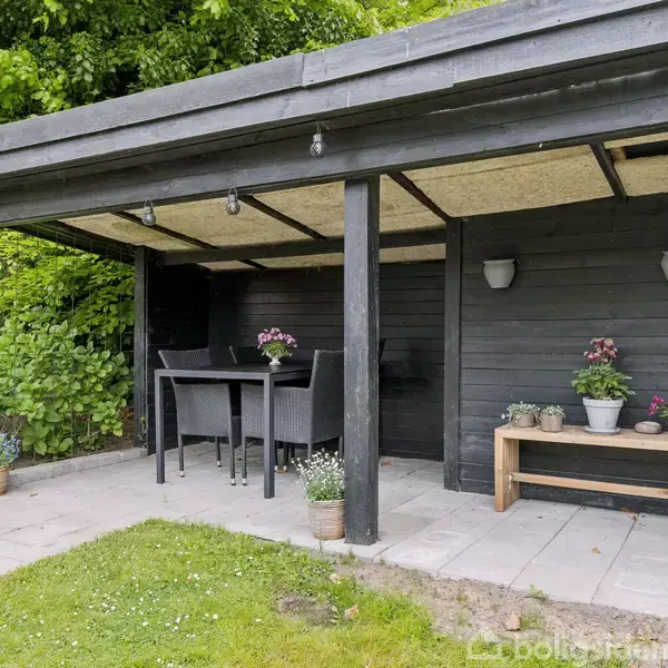 En sort træoverdækning beskytter et udendørs spisebord med stole. Blomsterkrukker står dekorativt på et bord. Omkring er der græsplæne og buskads, hvilket skaber en afslappende haveatmosfære.