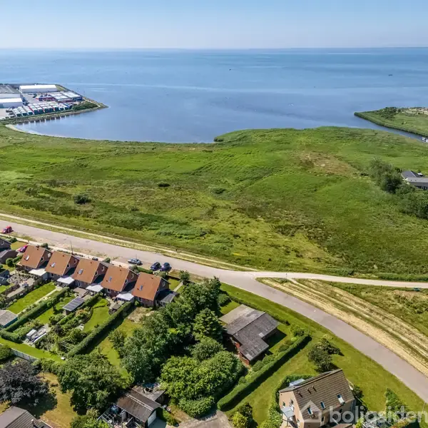 Rækkehuse med røde tage ligger langs en vej. Bag dem breder en grøn eng sig ud mod en fjord, hvor der ses et industriområde i baggrunden ved vandet.