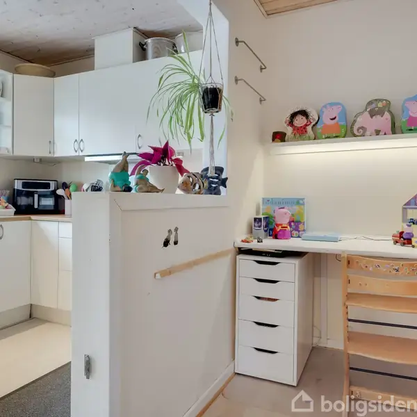 Et lyst køkken hjørne med hvide skabe og bordplader. Ved siden af er et børnearbejdsområde med en skrivebord, stol, legetøj og tegninger på hylden. Planter tilføjer dekoration.