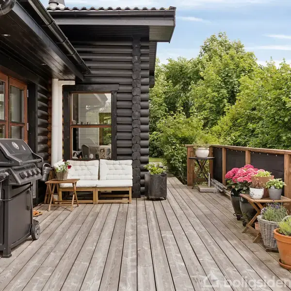 Grill på en træterrasse ved siden af et sort træhus. En hvid sofa med puder står ved huset. Planter og blomster i krukker pryder rækværket, omgivet af grønne træer.