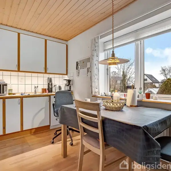 Et køkken med trægulv har et bord med stole ved et vindue. Loftet er i træ. Et skab og en mikroovn står ved væggen.