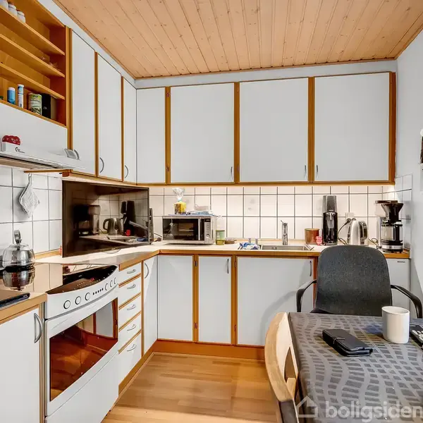 Et funktionelt køkken med hvidmalede skabe, trædetaljer og hvide fliser. En stol står ved et bord dækket med en grå dug. Elektriske apparater og køkkenredskaber er på bordpladerne.