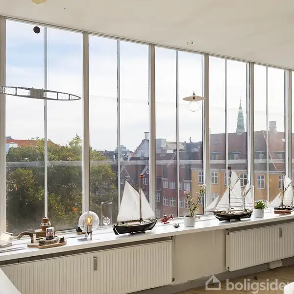 Modelskibe pryder vindueskarmen, stillet op foran store vinduer, der giver udsigt over byens tage og kirketårne. Der ses blomster og andre dekorative genstande i rummet.