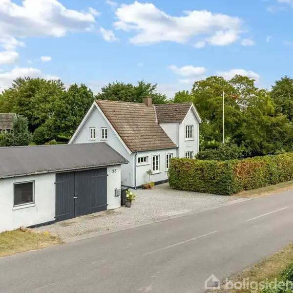 Et hvidt hus med brun tag står ved en vej, omgivet af træer og buske. Der er en garage til venstre og en adresseplade med nummeret "33".