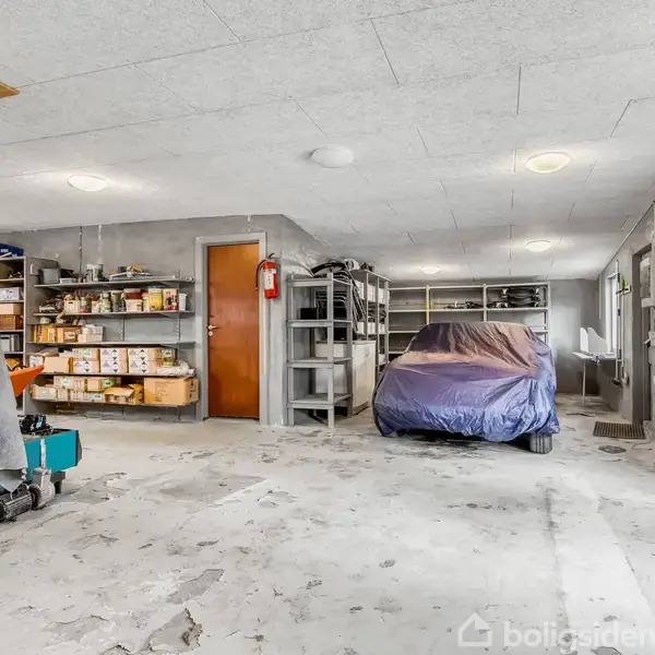 En garage indeholder en bil dækket med en presenning. Hylder langs væggen fyldt med kasser og værktøj. Gråt betongulv og lysloftlys. Dør ved bagvæggen.