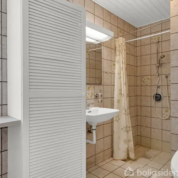 Et badeværelse med flisebelagte vægge og gulve; der er en håndvask, bruser med gardin samt et toilet. Et skab står ved siden af vinduet, og lyset er tændt over spejlet.