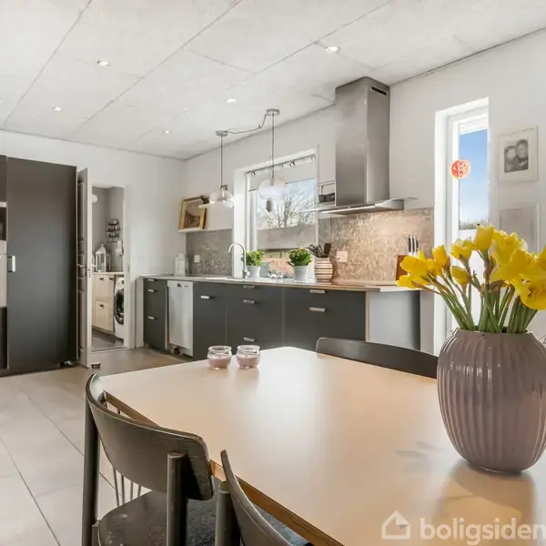 Bord med vase og gule blomster står i spisekøkken. Køkkenet har mørke skabe, indbygget ovn, og vinduer med naturligt lys. Stolene står ved bordet, under loftslamper.