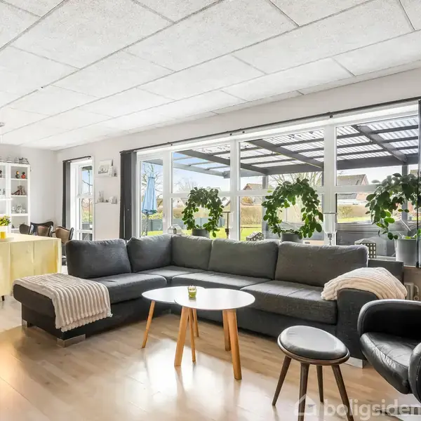 Stor grå sofa med tæpper, placeret foran runde sofaborde i en stue. Bagerst ses vinduer med udsigt til terrasse med planter, samt et spisebord med gul dug.