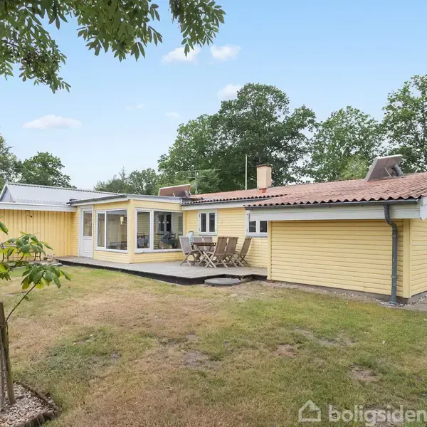 Et gult træhus med terrasse, omgivet af en græsplæne og træer. Et sæt havemøbler står på terrassen. Baggrunden er en skovdækket horisont under en blå himmel.