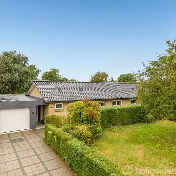 Et en-etages hus med gråt tag står i en have med grønne hække og træer. Til venstre er en garage og flisebelagt indkørsel.