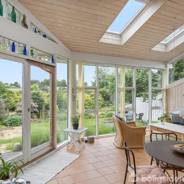Vinterhave med glasvægge og- loft. Rattanmøbler og spisebord står i et lyst rum, dekoreret med farvede glasflasker. Udsigt til en frodig have gennem vinduerne.