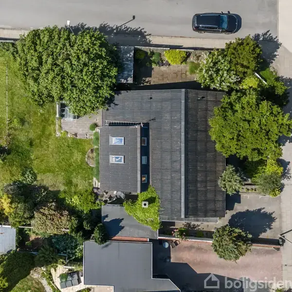 Et hus med sort tag ligger omgivet af en have med træer og græsplæne. Til venstre ses en vej med en bil parkeret.