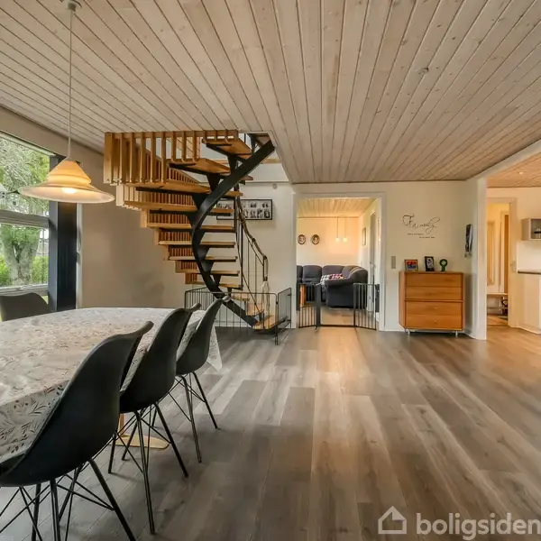 En elegant vindeltrappe af træ fører til øverste etage, i en stue med spisebord og stole, omgivet af store vinduer og trægulve med udsigt til en frodig have.