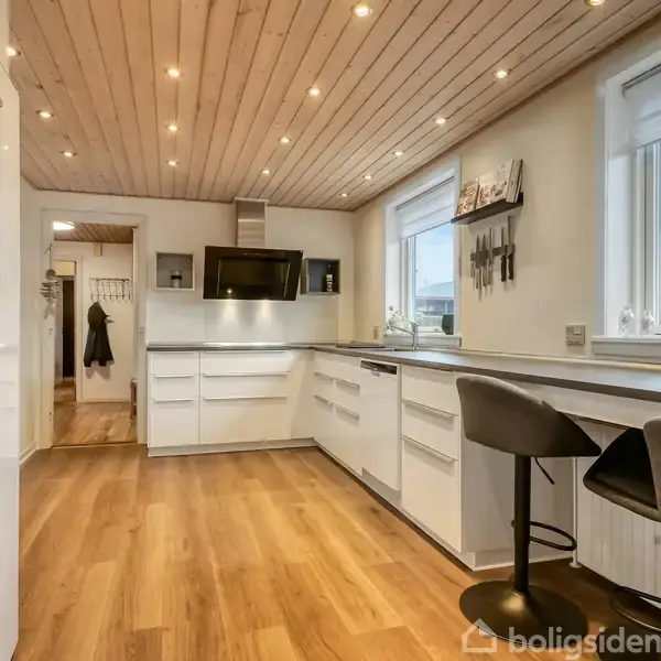 Et moderne køkken med hvide skabe, trægulv og træloft. Der er en bordplade med to barstole ved vinduet, vægmonteret emhætte og indbygget ovn.