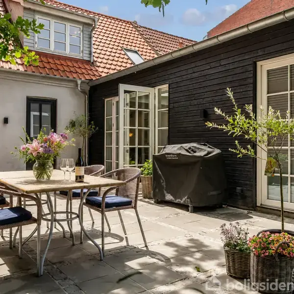 Udendørs gårdhave med et bord og fire stole. Bordet er dekoreret med blomster. En grill står tæt ved sorte bræddevægge. Sollys kaster skygger på flisebelægningen.