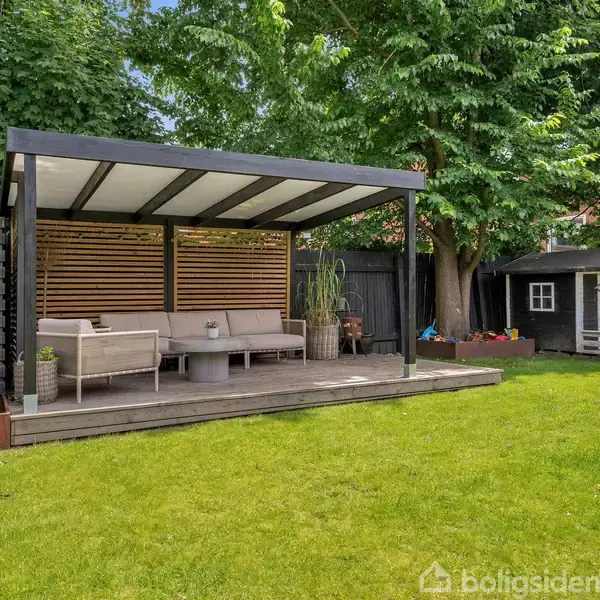 Overdækket terrasse med sofa og bord står på en træplatform i en grøn have. Ved siden af står et legehus i træ. Store træer omkranser området.