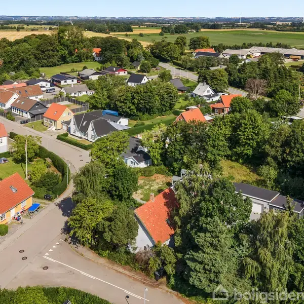 Boliger med røde og mørke tage står stille i et grønt naturområde. Huse er omgivet af træer og marker, hvilket skaber en rolig forstadsmiljø.