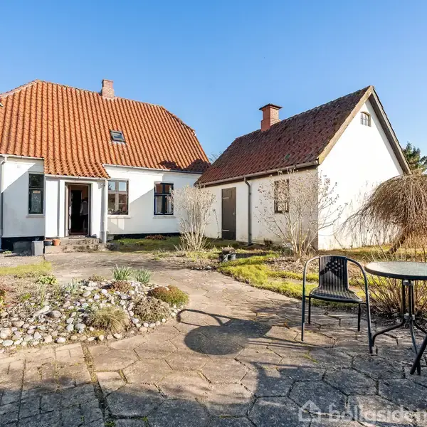 Et hvidt hus med rødt tegltag står i en solrig have. Haven har en flisebelagt terrasse med bord og stole, omgivet af buske og en lille stenhave.