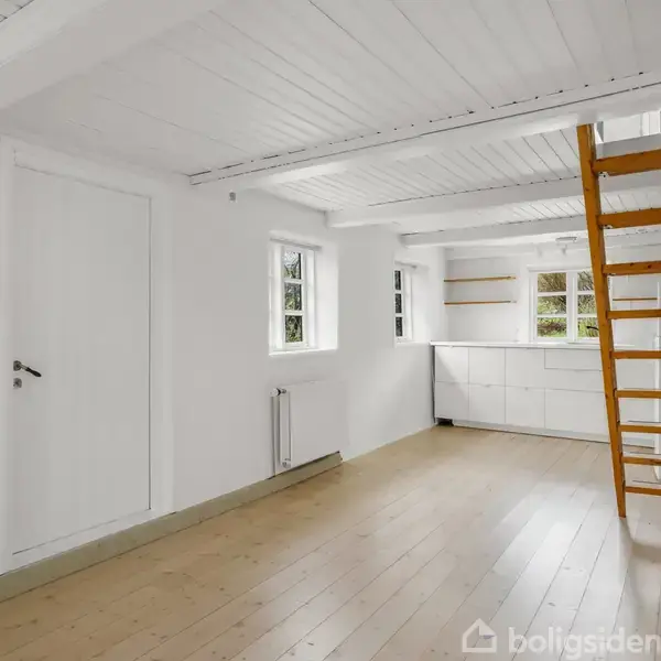 Et lyst rum med hvidmalede vægge og træloft. Trætrappe fører opad. To små vinduer og en radiator findes på væggene. Rummet fremstår minimalistisk og uindrettet.