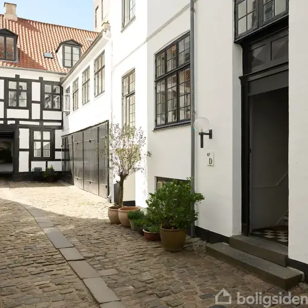 En gård med brostensbelægning og hvide bygninger, dørmærket "D". Der står planter i potter langs væggen; det er en rolig og velplejet atmosfære.