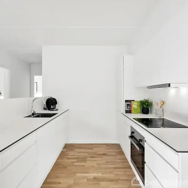Et moderne, lyst køkken med hvide skabe. En kogeplade og ovn er integreret i højre side, mens en vask og kaffemaskine er på venstre side. En vase med blomster tilføjer farve.