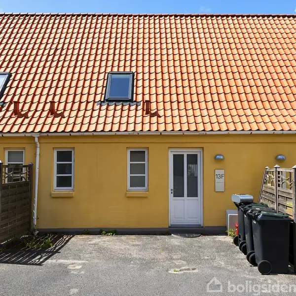 Et gult rækkehus med rødt tegltag, tre tagvinduer, og en hvid dør markeret "13F". Affaldscontainere står ved siden, omkranset af træhegn. Asfalteret parkeringsplads foran.