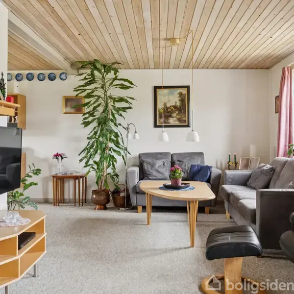 Stue med sofaarrangement ved et hjørnebord, omgivet af planter og dekorationer. Træbeklædt loft og tæppebelagt gulv. Sofaen er mod et vindue med røde gardiner, som giver naturligt lys.