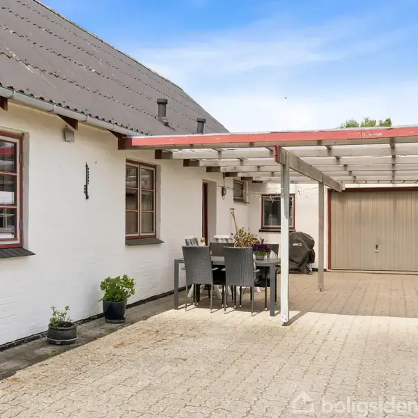 Hus med hvid facade, sort tag, røde vinduer og en overdækket terrasse med spisebordssæt. Terrassen har en garage ved siden af. Blomsterkrukker står langs væggen.