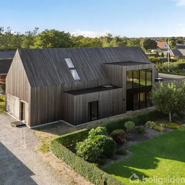 Et moderne træhus med asymmetrisk tag ligger centralt i en velplejet have med buske og græsplæner, omgivet af andre huse.