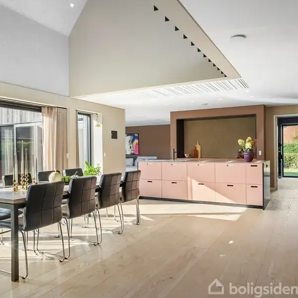 Spisebord med sorte stole står i et lyst, moderne køkken-alrum. Store vinduer til venstre giver udsigt til haven. Køkkenelementer og planter bidrager til en indbydende atmosfære.