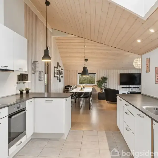 Et moderne køkken med hvide skabe og grå bordplader. I baggrunden er en spisestue med belysning og vindue. Loftet er træbeklædt, skaber en lys atmosfære.
