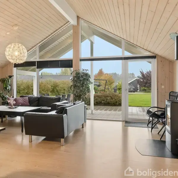 Stue med sorte sofaer og brændeovn. Store vinduer giver udsigt til have. Moderne indretning inkluderer spisebord og fladskærms-tv. Lampe hænger fra højt, skrånende træloft.