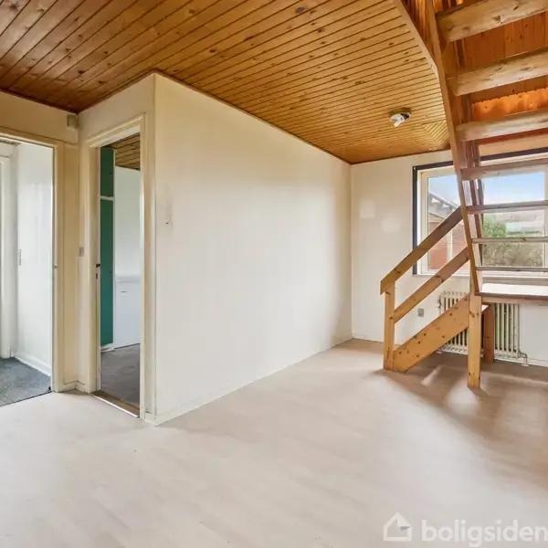 Et rum med en trætrappe, der fører til en øvre etage. Der er lyse trægulve og træloft. En dør til venstre fører ud til en entre med fliser.