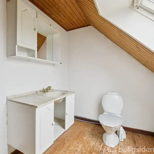 Håndvask med spejl hænger ved væg, tæt på et toilet. Loftet er skråt med træpaneler. Trægulve og en skrå rude skaber et lyst rum. Ingen tekst til stede.