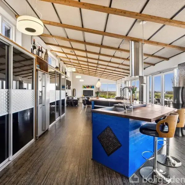 Køkken med moderne indretning, inkluderer en stor blå køkkenø og barstole, med udsigt gennem store vinduer til en grøn have. Loftet har træbjælker og indbyggede lamper.