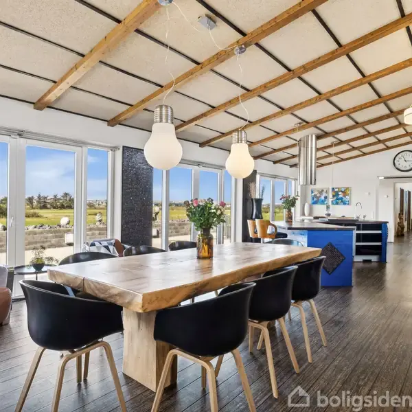 Et stort træbord med sorte stole står i en moderne spisestue. Store vinduer giver udsigt til en grøn have. Lysekroner hænger fra et træbeklædt loft.