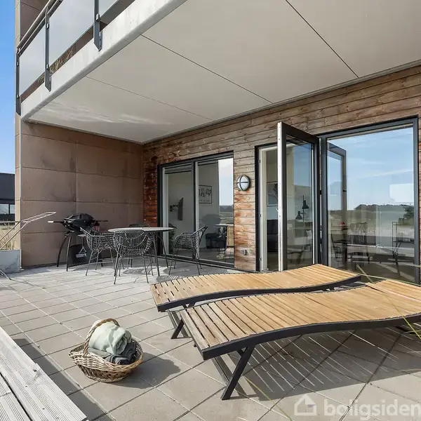 To liggestole står på en flisebelagt terrasse foran en bygning med træpaneler. En kurv med et tæppe ligger ved siden af. I baggrunden er bord og stole samt en grill.