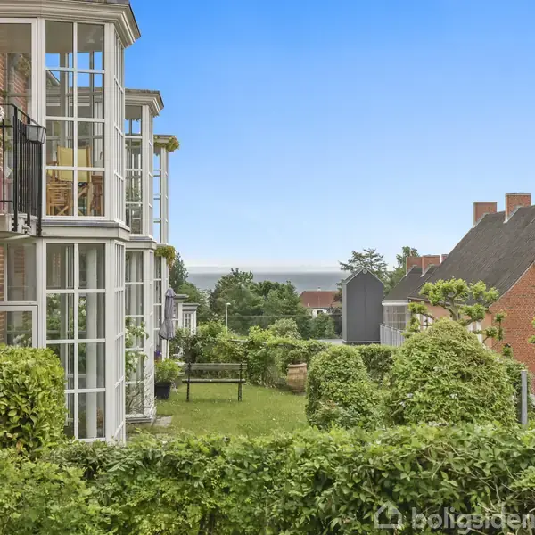 En række altaner med glas og blomsterkasser strækker sig ud fra en murstensbygning. De er omgivet af velplejede hække, med udsigt til havet og andre bygninger i baggrunden.
