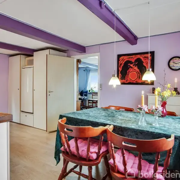 Et spisebord med grøn dug og fire træstole står i et lilla malet køkken med fritlagte bjælker. Over bordet hænger lamper, mens blomster og stearinlys dekorerer bordet.